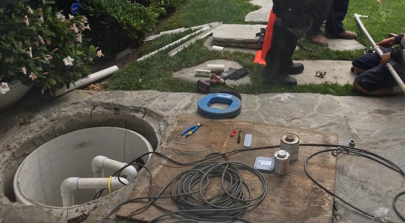 Open septic pump chamber in a stone patio with wiring and PVC primer on a plywood board
